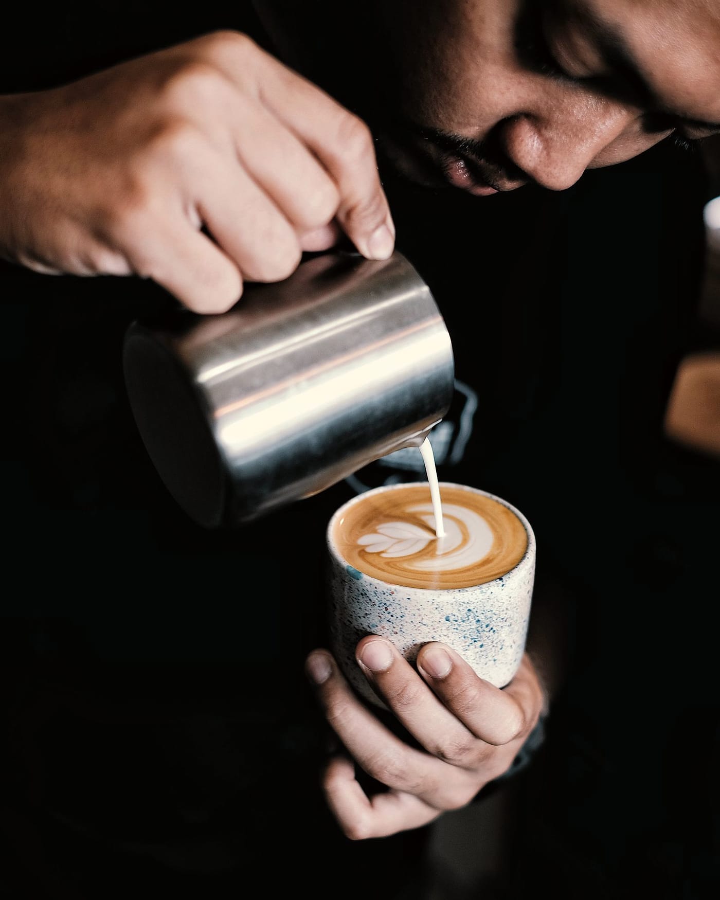 A barista pours steamed milk into a speckled cup held low in one hand.