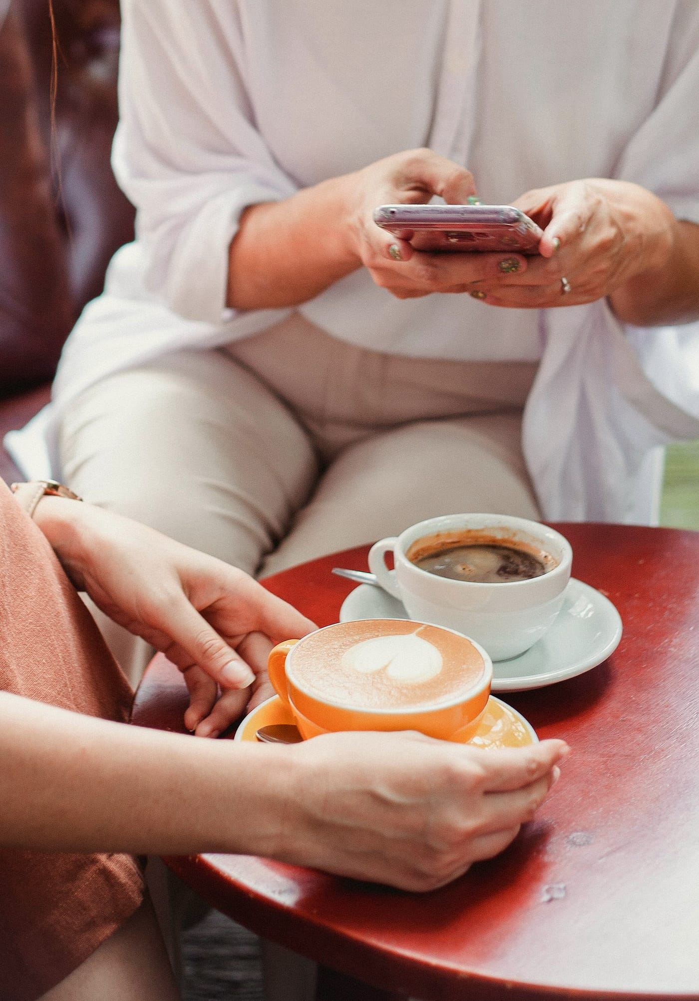 Two friends at a red table, one on her phone, coffee cups between them.